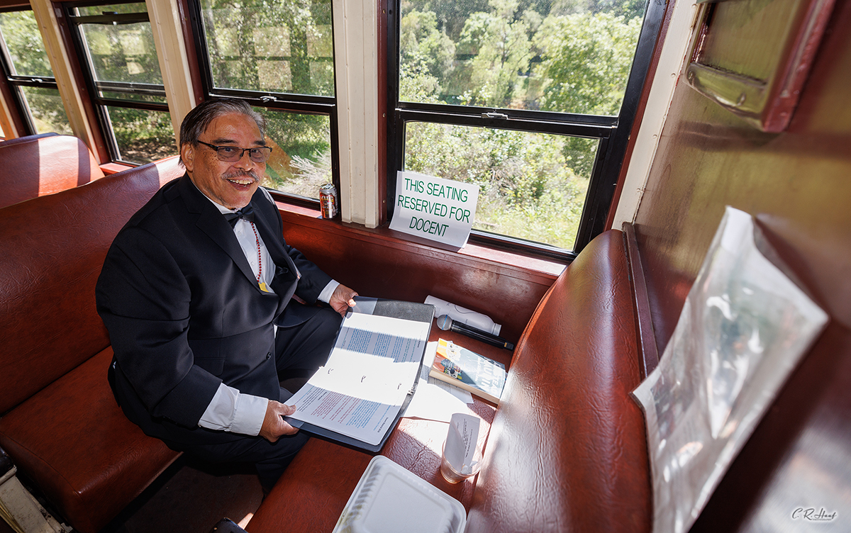 A man in a suit and bowtie is seated inside a historic railroad car with a notebook sitting on the seat in front of him that contains the different historical items to share as the docent on the train.